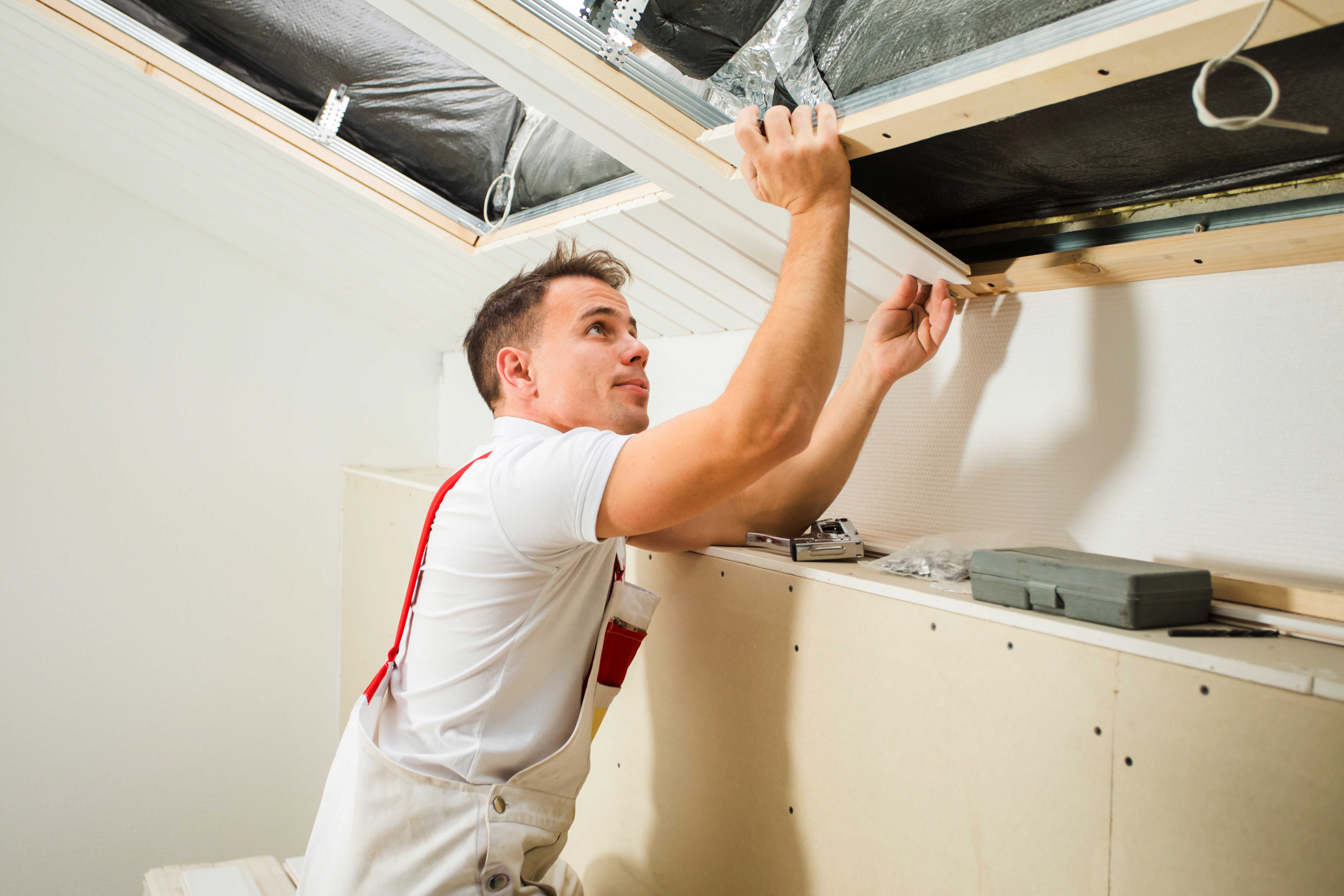 Håndværker iført hvid trøje og arbejdsbukser arbejder på montering eller reparation af et skråloft. Han bruger begge hænder til at justere en træramme, mens værktøj og materialer ligger på en arbejdsflade ved siden af.