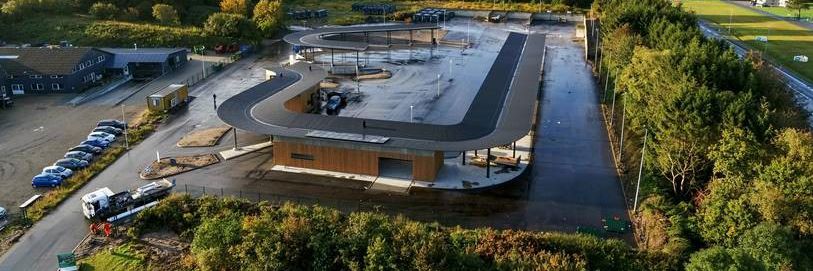 Luftfoto af Give Genbrugsplads omgivet af grønne områder og træer. Pladsen har en tydelig cirkulær rute for biler og adskillige containere og stationer til affaldssortering. Bygninger og overdækkede områder sikrer, at besøgende nemt kan navigere gennem genbrugspladsen.