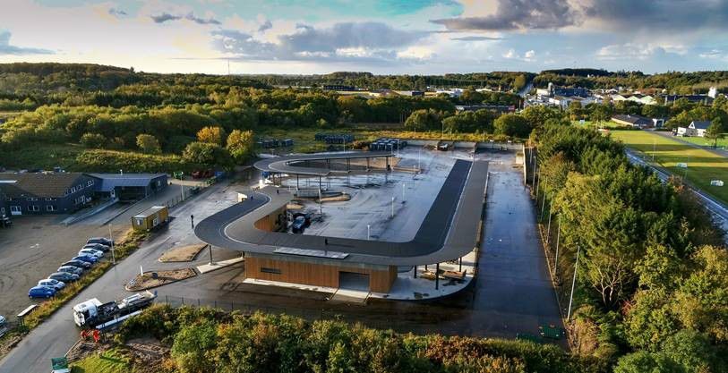 Luftfoto af Give Genbrugsplads omgivet af grønne områder og træer. Pladsen har en tydelig cirkulær rute for biler og adskillige containere og stationer til affaldssortering. Bygninger og overdækkede områder sikrer, at besøgende nemt kan navigere gennem genbrugspladsen.