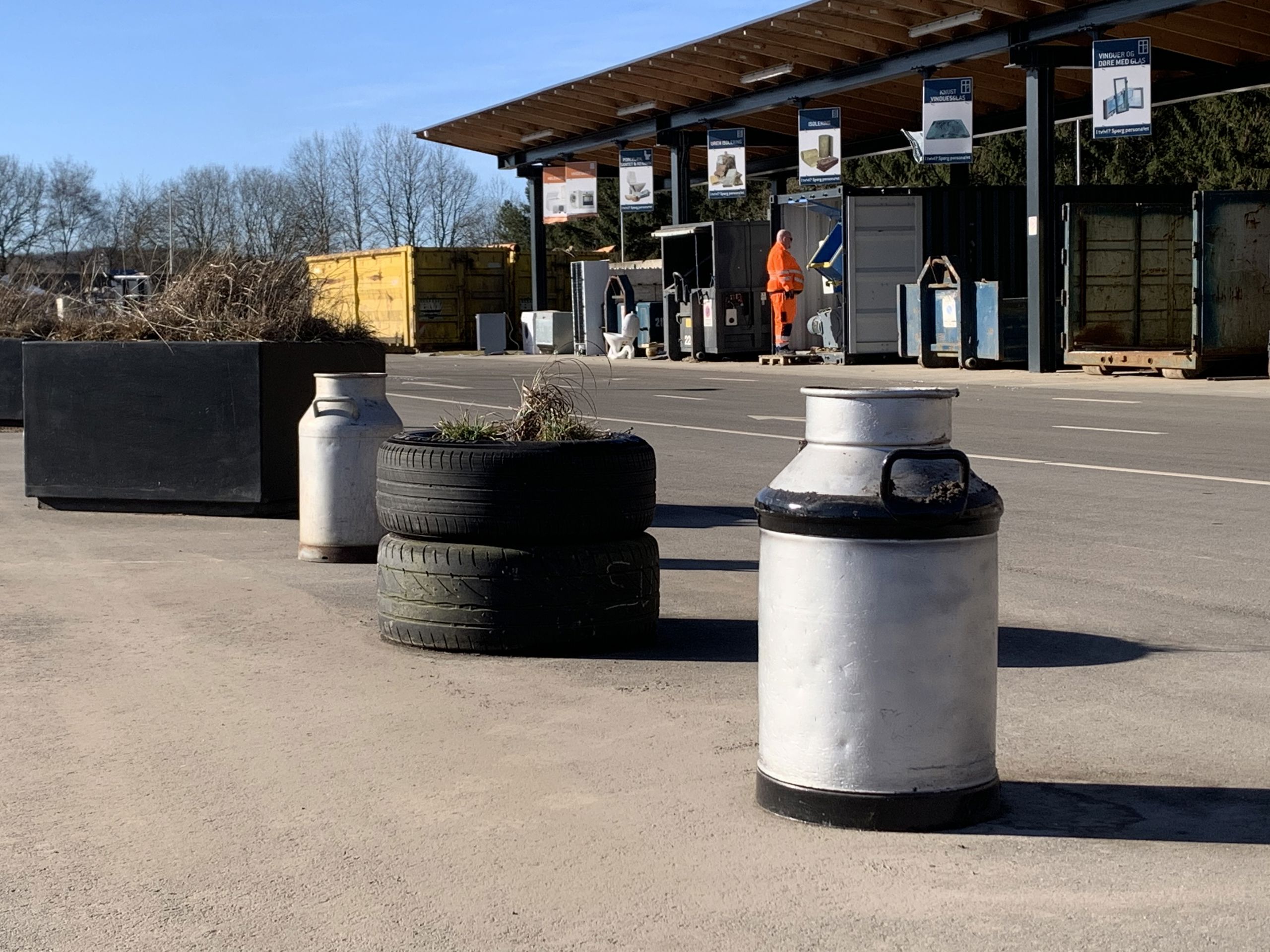 Foto fra Give Genbrugsplads med gamle mælkejunger og dæk brugt som dekoration ved siden af affaldsstationerne. I baggrunden ses containere og en medarbejder i arbejdstøj, som guider besøgende i affaldssortering. Skilte hænger fra taget og viser, hvor forskellige typer affald kan afleveres.