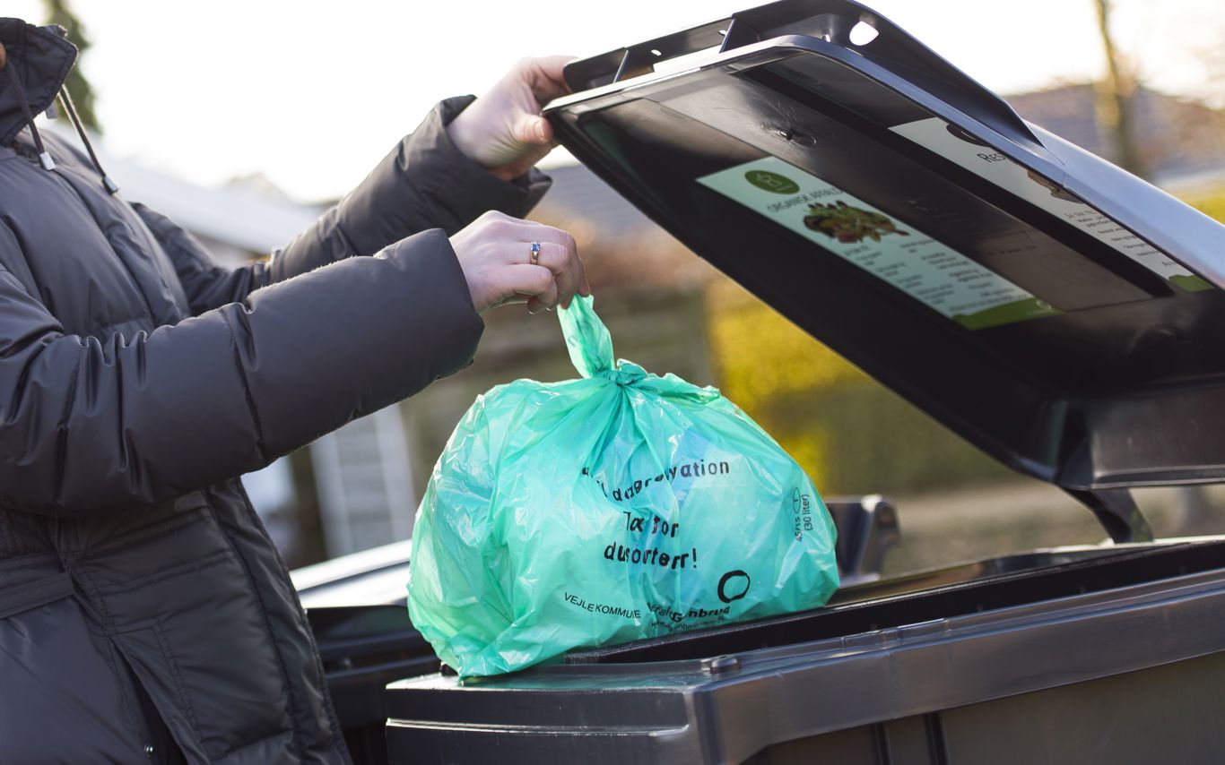erson iført mørk jakke smider en grøn biologisk nedbrydelig pose med madaffald i en sort affaldscontainer. Containerlåget har en informationsplakat om korrekt affaldssortering. Baggrunden viser en villavej med hække og huse i sollys.