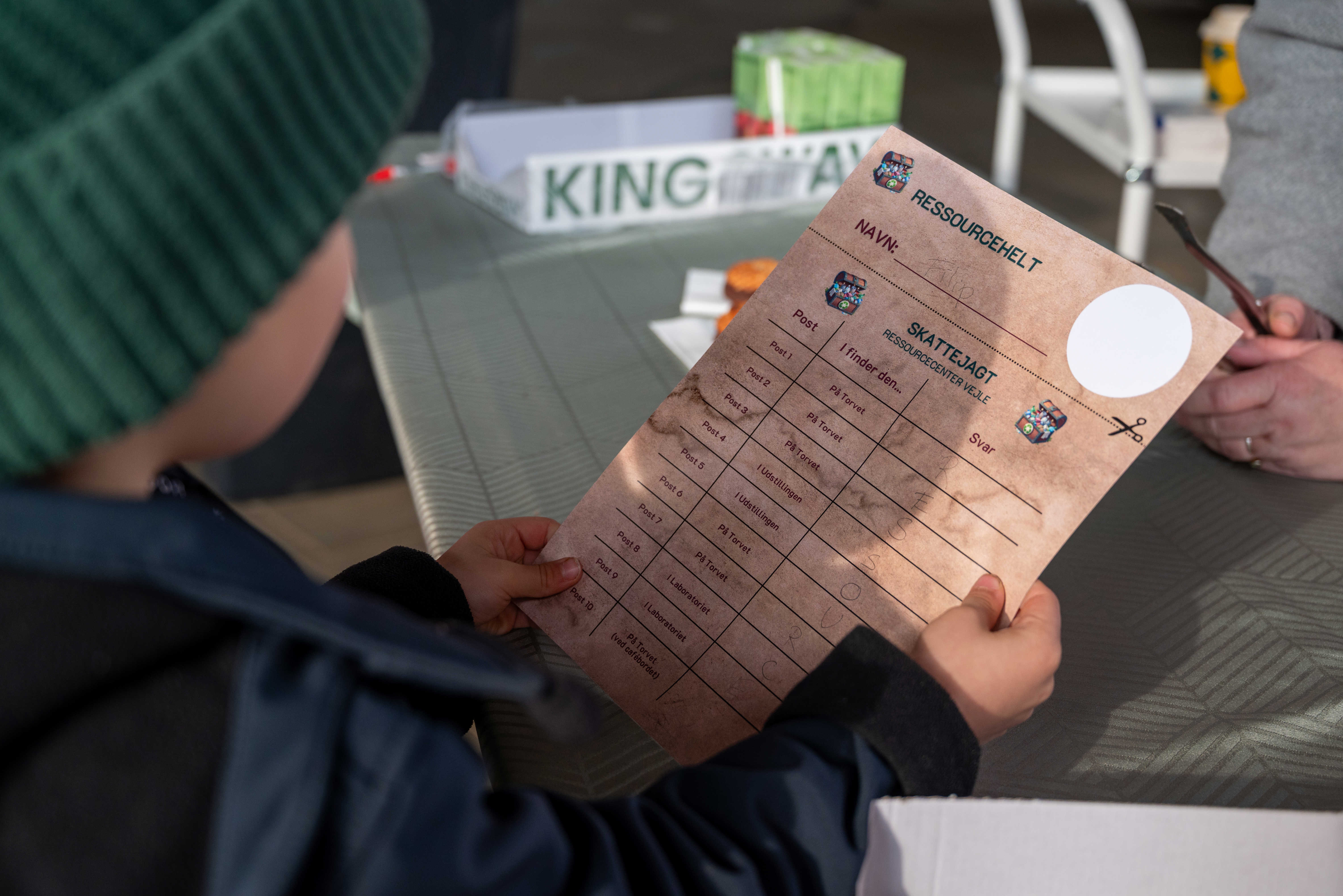 Barn står med papir i hånden, hvorpå der står "ressourcehelt" som en del af skattejagt på Torvet ved RessourceCenter Vejle i forbindelsen med fejringen af RCV's 2-års fødselsdag-