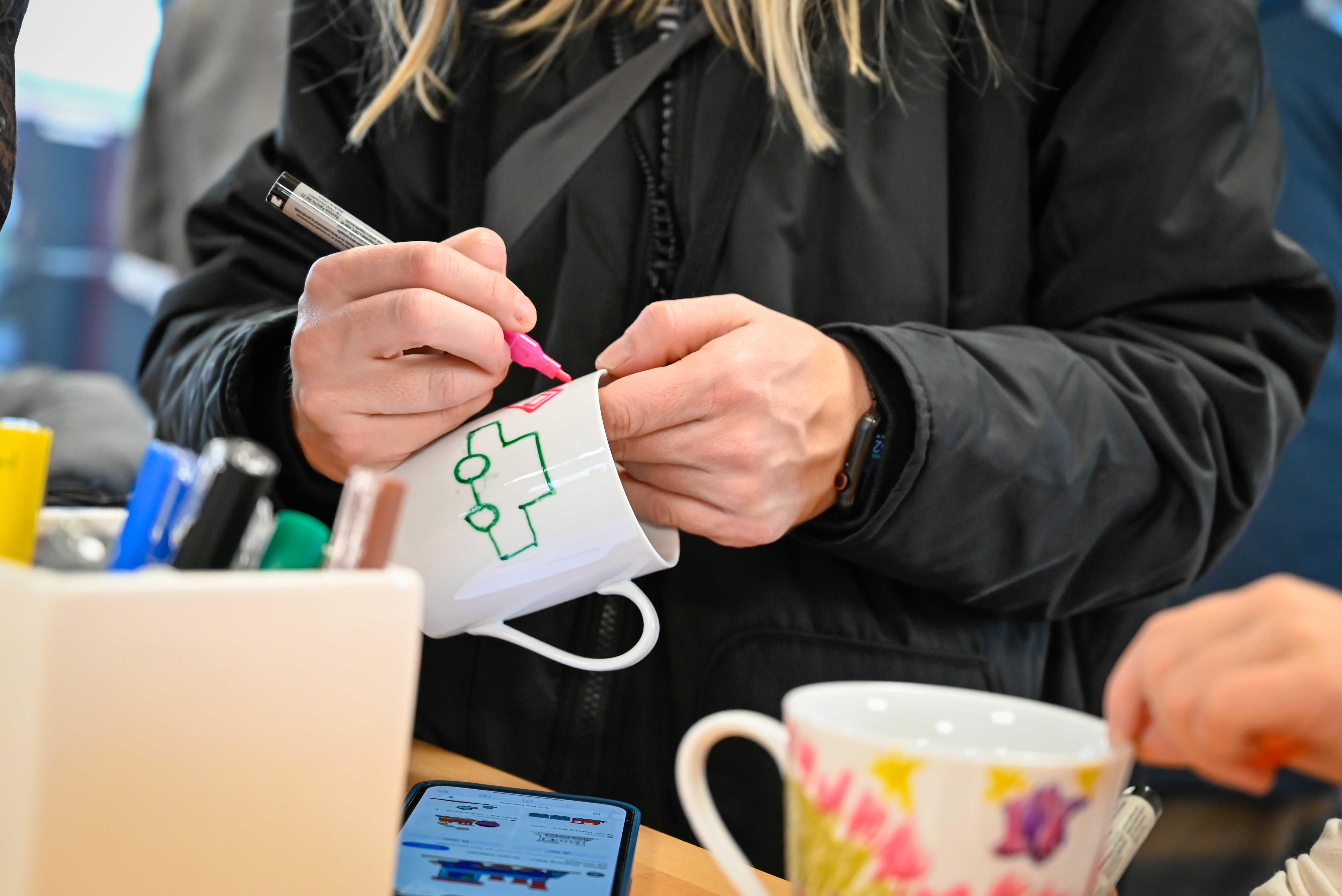 Person iført sort jakke maler med tusch på hvid kop af keramik.