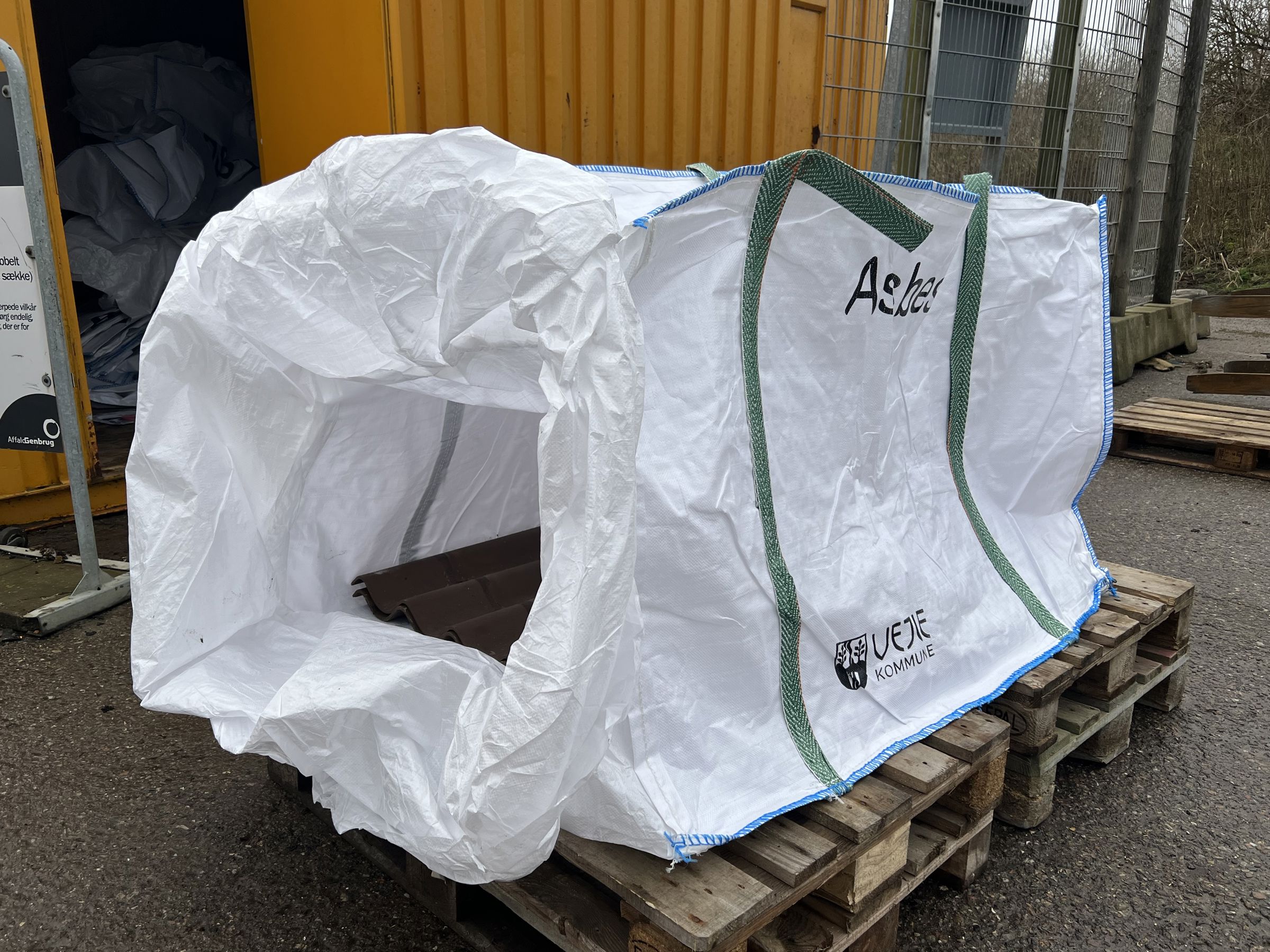 Stor hvid asbest-sæk med grønne håndtag og Vejle Kommunes logo, placeret på en træpalle ved siden af en gul container. Sækken er åben og klar til brug