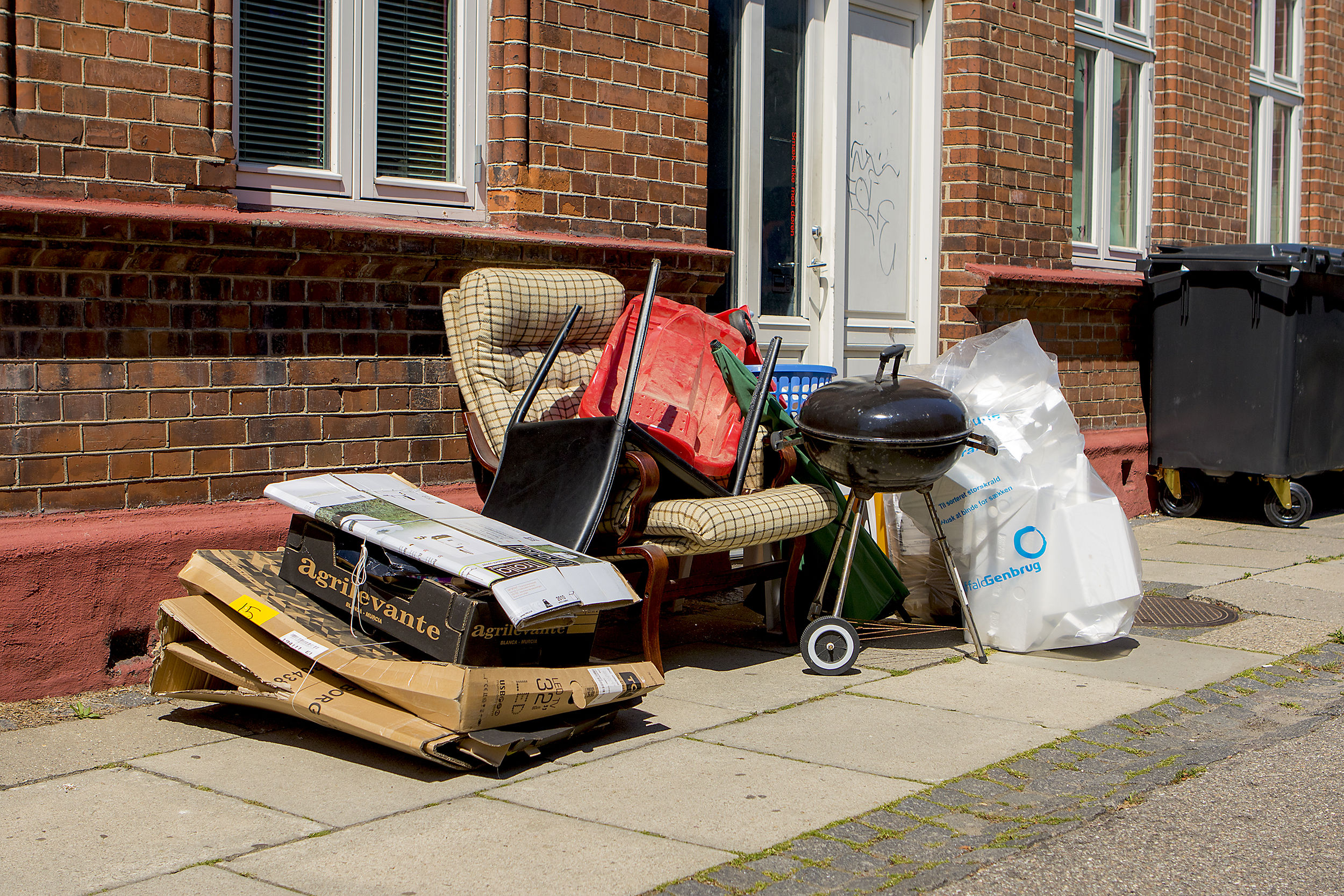 torskrald sat ud på fortovet foran en murstensbygning. En lænestol, papkasser, en grill, en sæk med affald og diverse genstande venter på afhentning. Sorte affaldscontainere står i baggrunden ved husmuren.