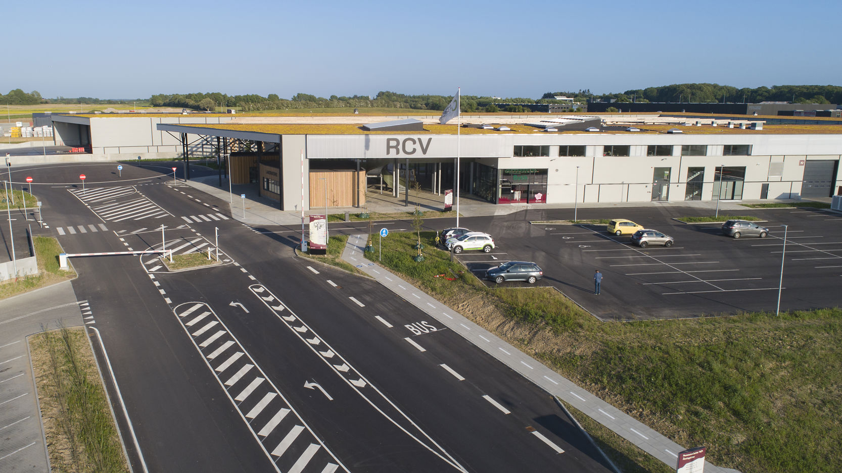 Luftfoto af RessourceCenter Vejle (RCV). Centret har en stor bygning med fladt tag, en parkeringsplads og tydeligt markerede vejbaner og indkørsler.
