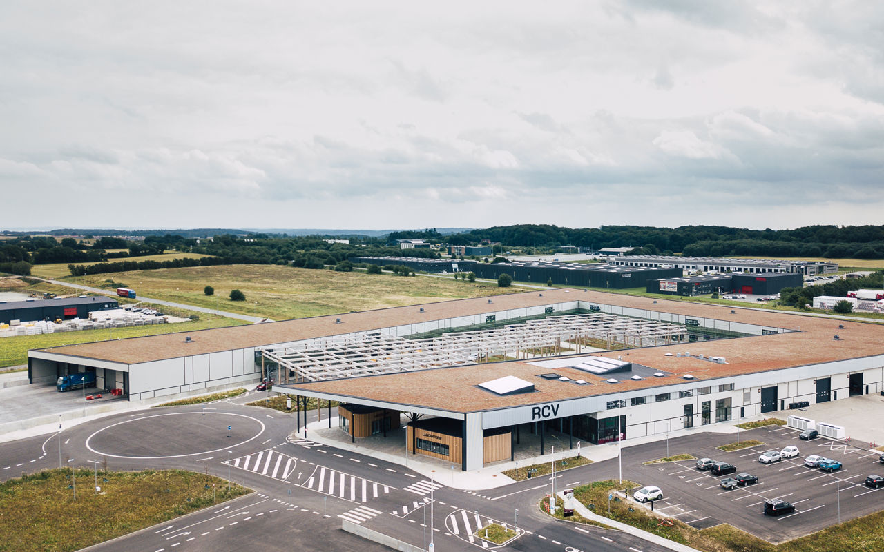 Luftfoto af RessourceCenter Vejle (RCV). Centret har en stor bygning med fladt tag, en parkeringsplads og tydeligt markerede vejbaner og indkørsler. Taget har solpaneler, og der er en åben konstruktion med trærammer i gårdområdet.