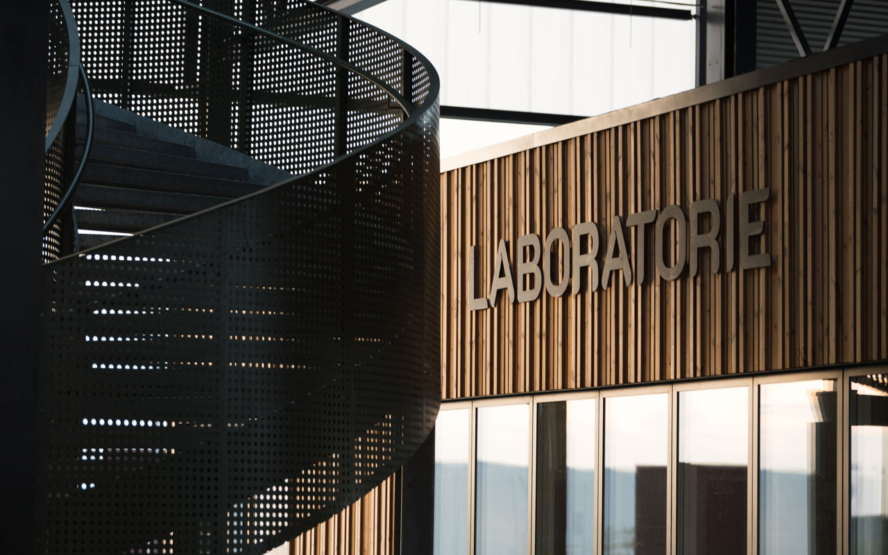 Nærbillede af facaden til Laboratoriet på RessourceCenter Vejle (RCV). Facaden er lavet af trælameller, og ordet "LABORATORIE" er monteret i store bogstaver. Til venstre ses en moderne vindeltrappe i perforeret metal, der tilføjer et industrielt præg til designet.