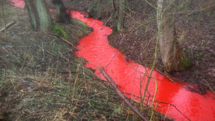 Rød maling hældt i en vejrist, som er endt i et vandløb.