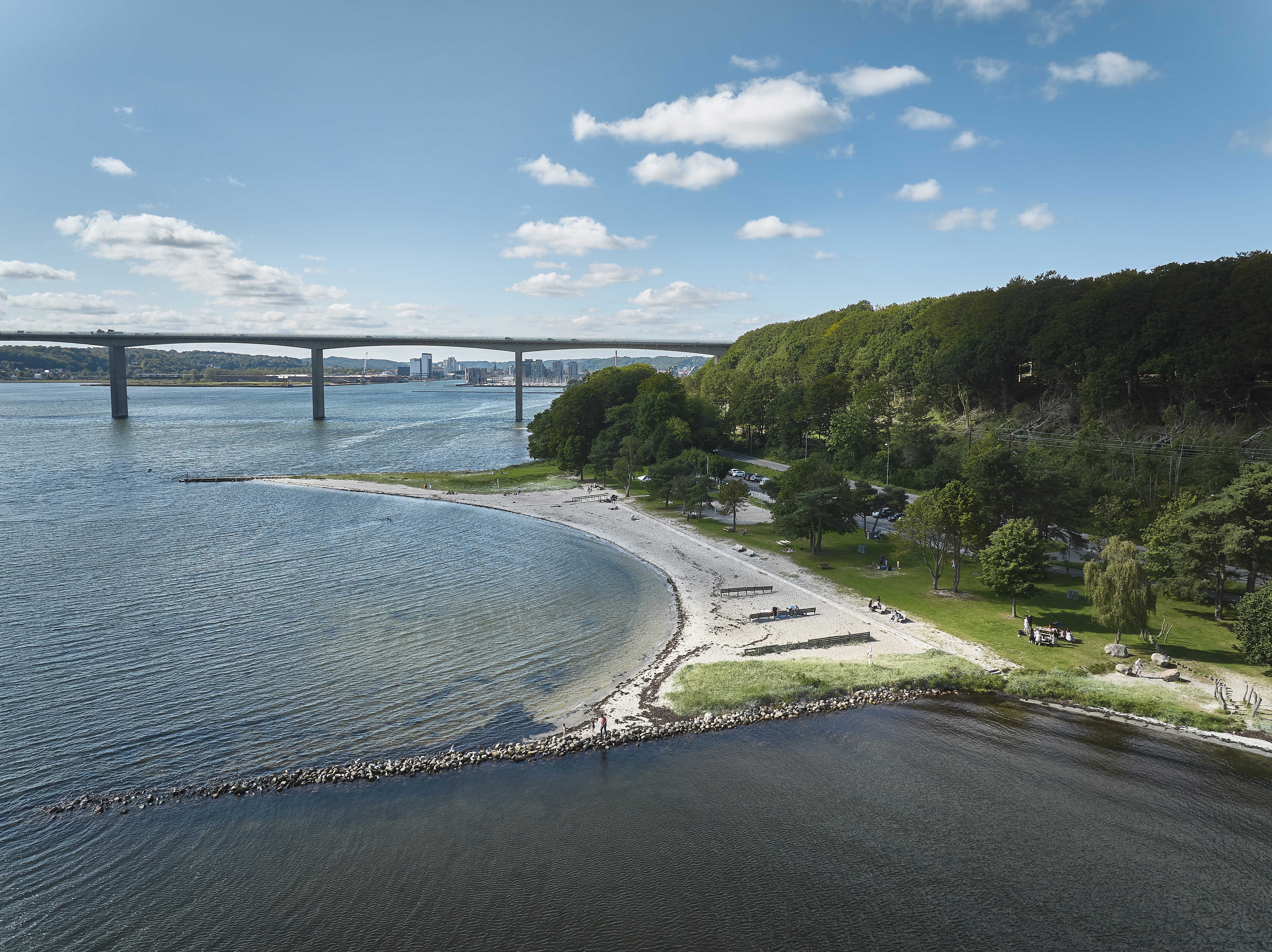 Kystlandskab med en sandstrand og grønne arealer langs vandet. I baggrunden ses Vejlefjordbroen, der strækker sig over fjorden med byen Vejle i horisonten.