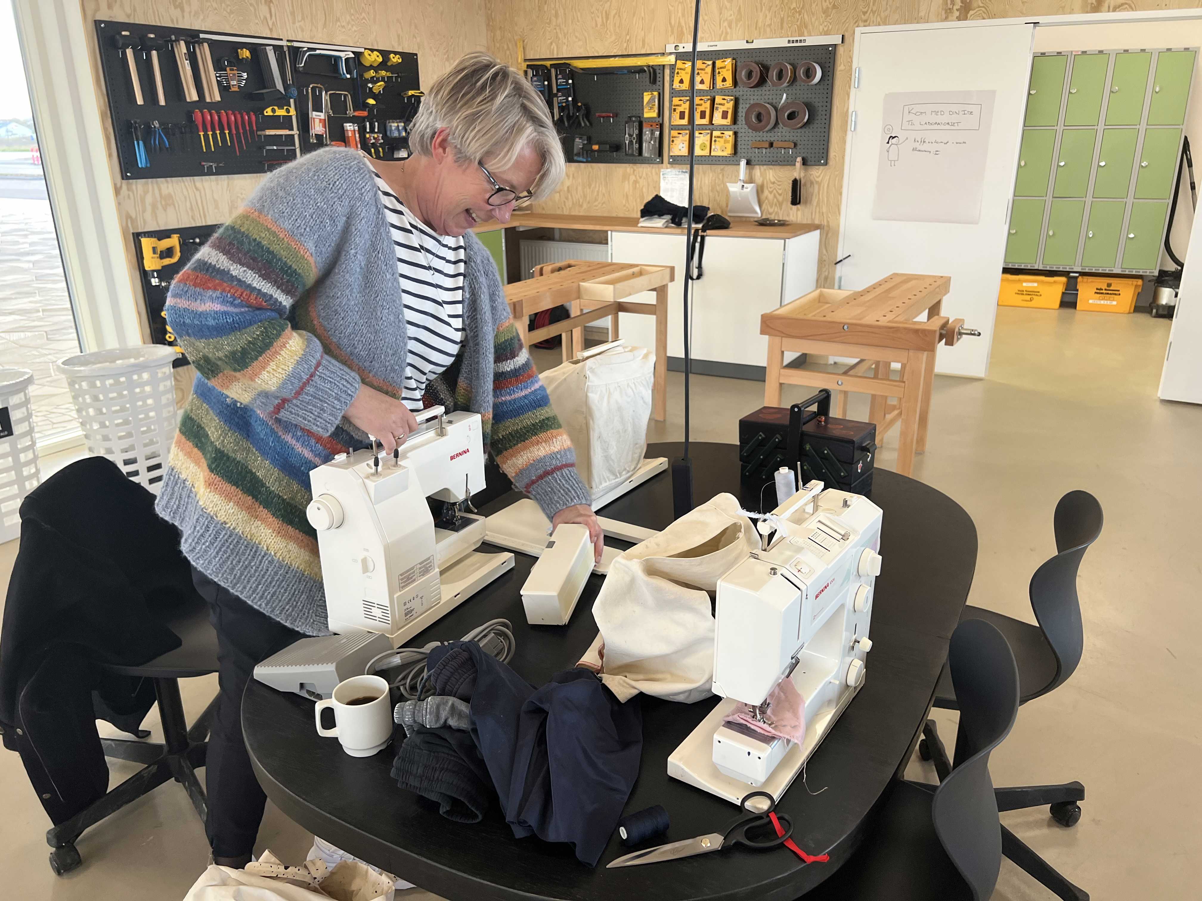 Smilende kvinde ved symaskine i Laboratoriet på RCV. I baggrunden ses træværkstedshjørnet med slagbænke og værktøj.