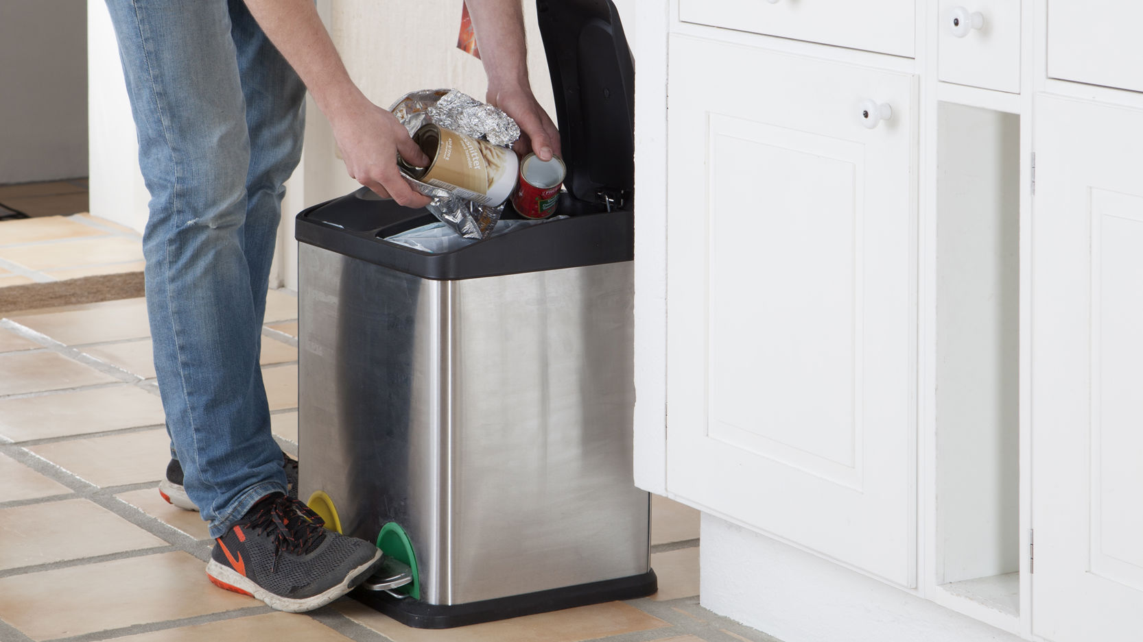 Person i jeans og sneakers smider affald i en skraldespand af rustfrit stål i et køkken med hvide skabe og flisegulv.