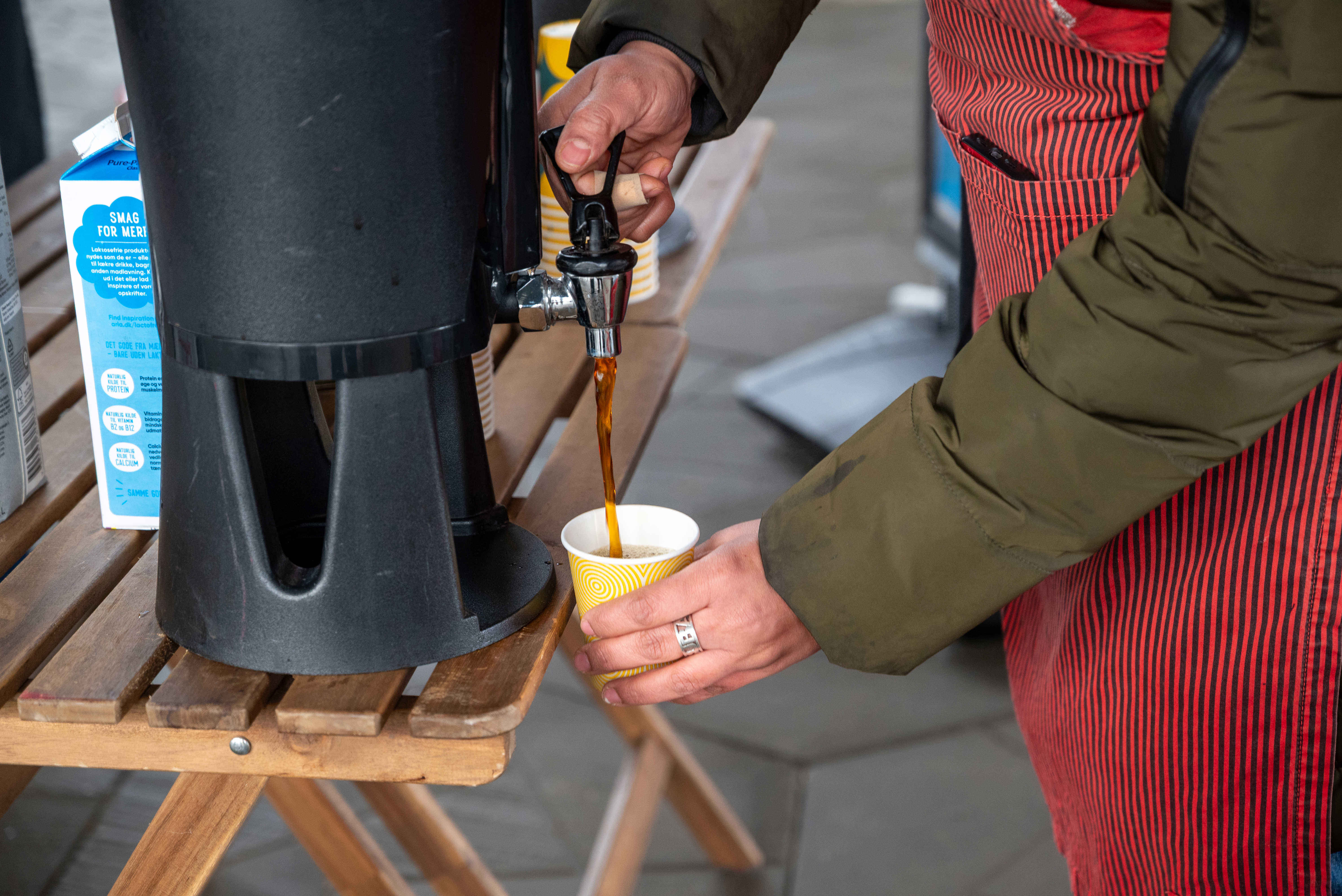 Person iført grøn jakke og rødt forklæde hælder kaffe i et krus på Torvet ved RessourceCenter Vejle i forbindelsen med fejringen af RCV's 2-års fødselsdag-