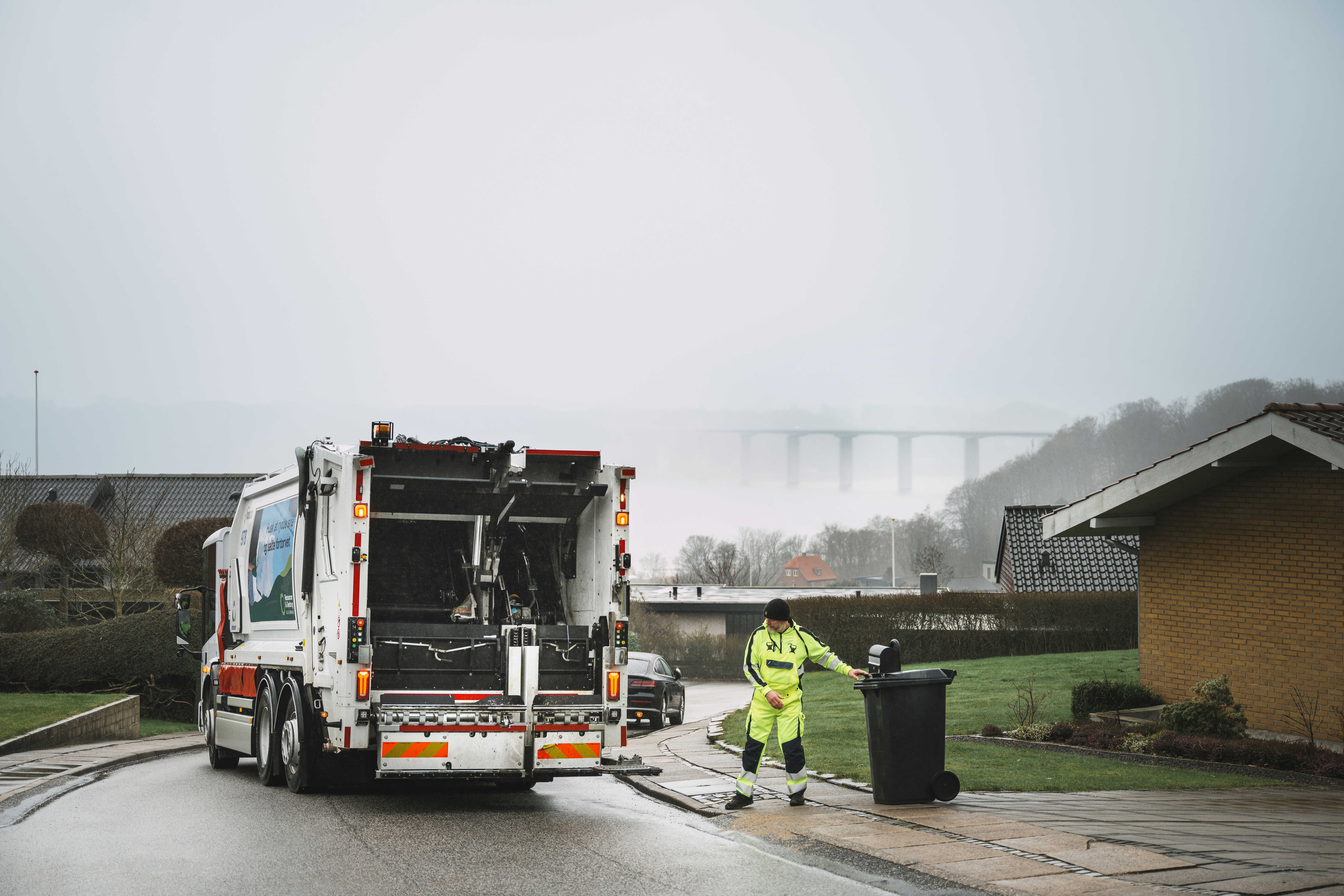 Bagenden af en skraldebil under arbejdet, med udsigt over et boligområde.