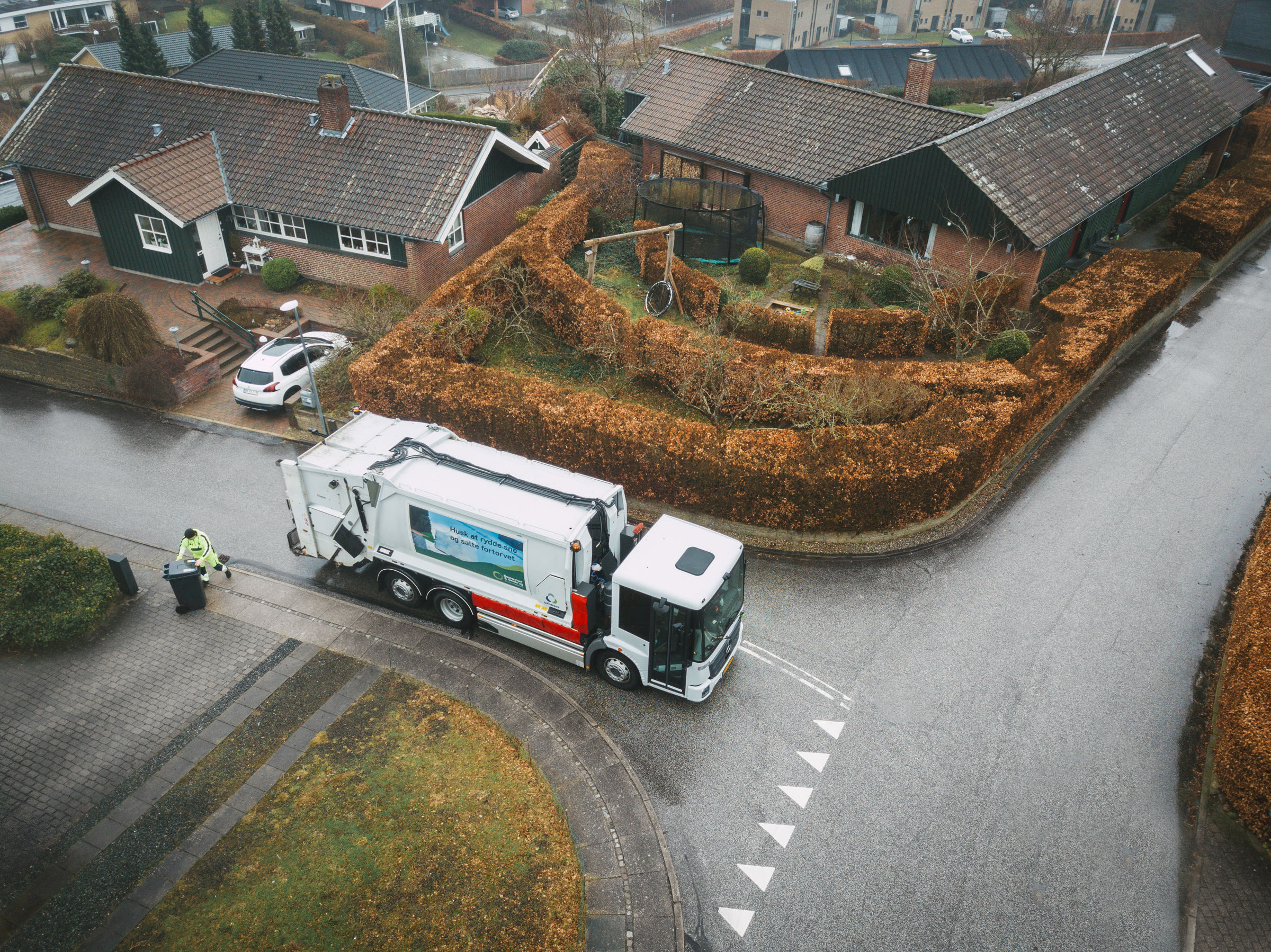 En skraldebil, der kører rundt i et boligområde med velplejede hække, mens en skraldemand tømmer en affaldsbeholder