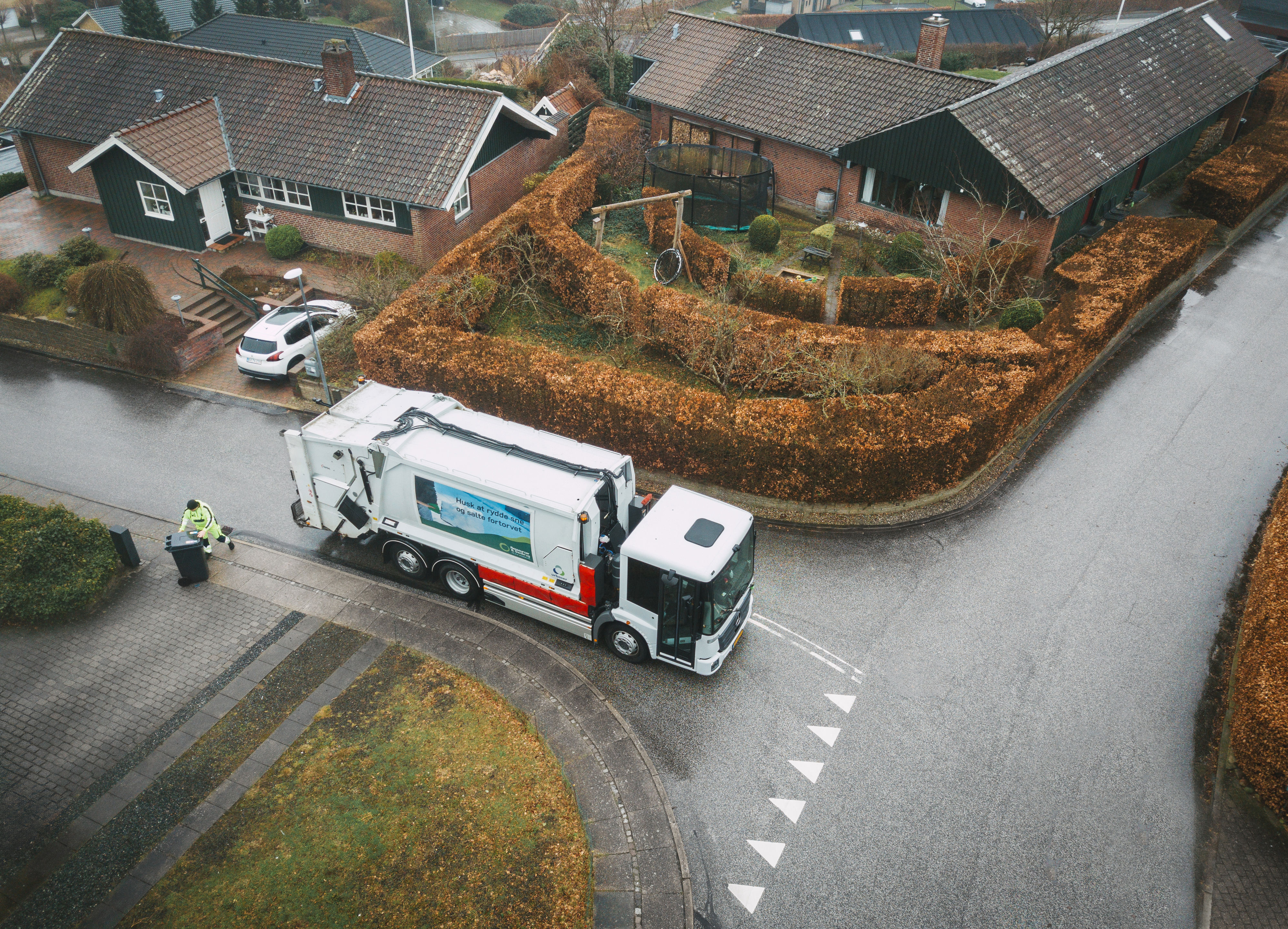 En skraldebil, der kører rundt i et boligområde med velplejede hække, mens en skraldemand tømmer en affaldsbeholder