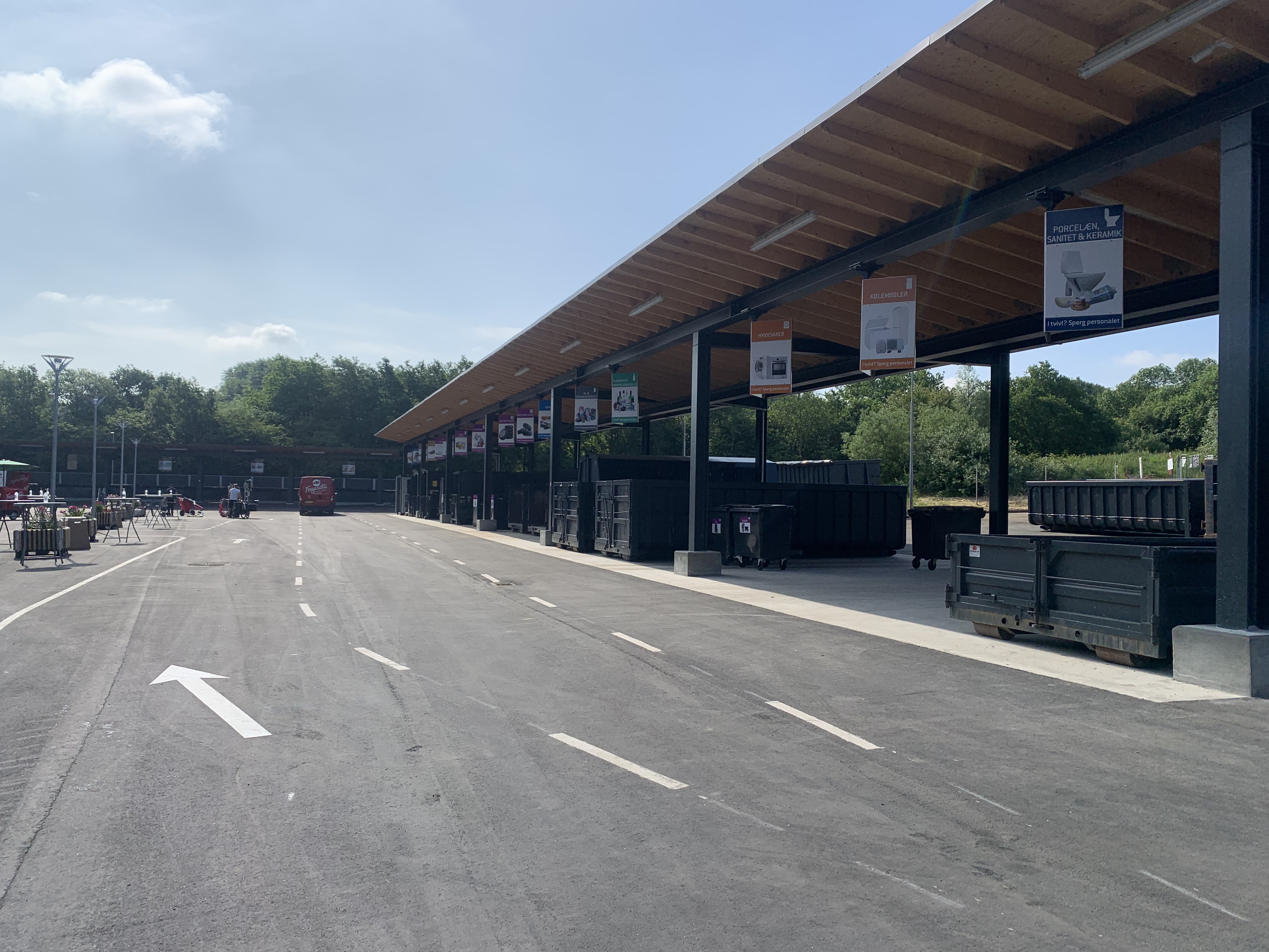 Børkop Genbrugsplads med en lang overdækket affaldssorteringsstation. Store containere er placeret under skiltede sektioner for forskellige affaldstyper. En asfalteret vej med kørselsanvisninger fører gennem pladsen, hvor en rød varevogn er synlig i baggrunden. Omkring pladsen er der grønne træer og en blå himmel med få skyer.