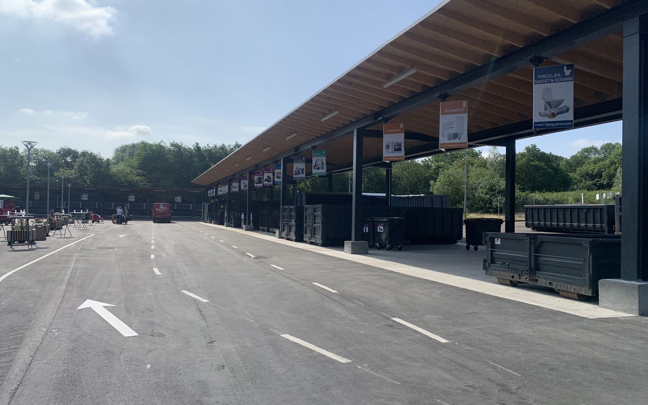 Børkop Genbrugsplads med en lang overdækket affaldssorteringsstation. Store containere er placeret under skiltede sektioner for forskellige affaldstyper. En asfalteret vej med kørselsanvisninger fører gennem pladsen, hvor en rød varevogn er synlig i baggrunden. Omkring pladsen er der grønne træer og en blå himmel med få skyer.