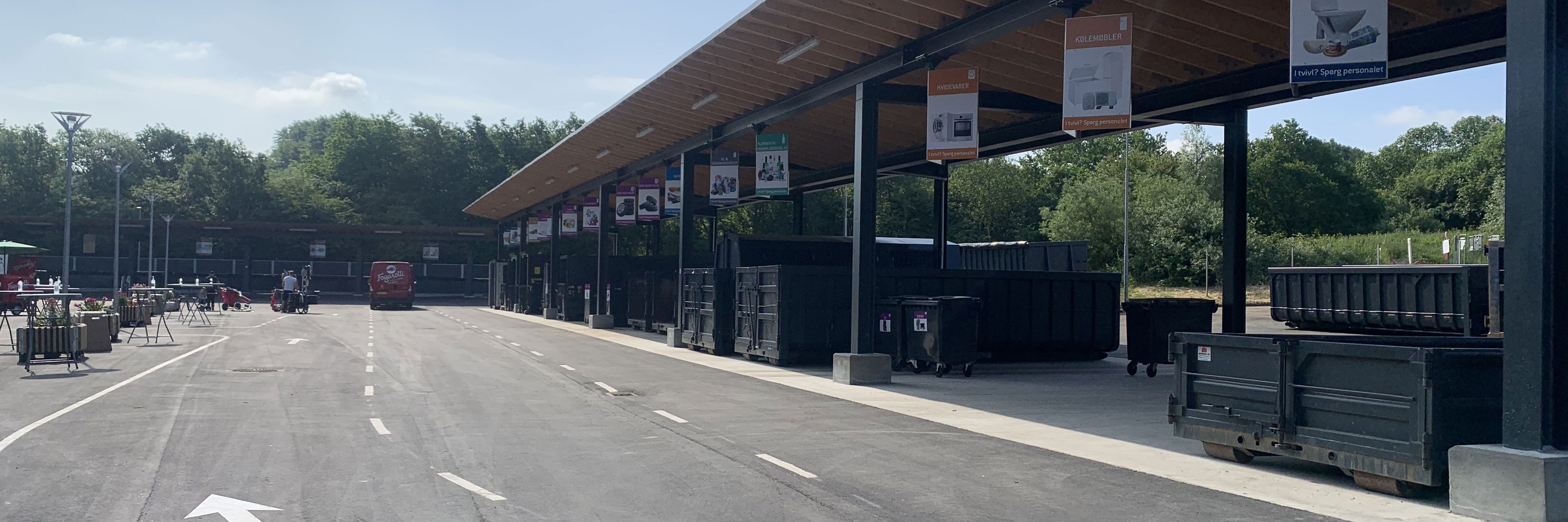 Børkop Genbrugsplads med en lang overdækket affaldssorteringsstation. Store containere er placeret under skiltede sektioner for forskellige affaldstyper. En asfalteret vej med kørselsanvisninger fører gennem pladsen, hvor en rød varevogn er synlig i baggrunden. Omkring pladsen er der grønne træer og en blå himmel med få skyer.