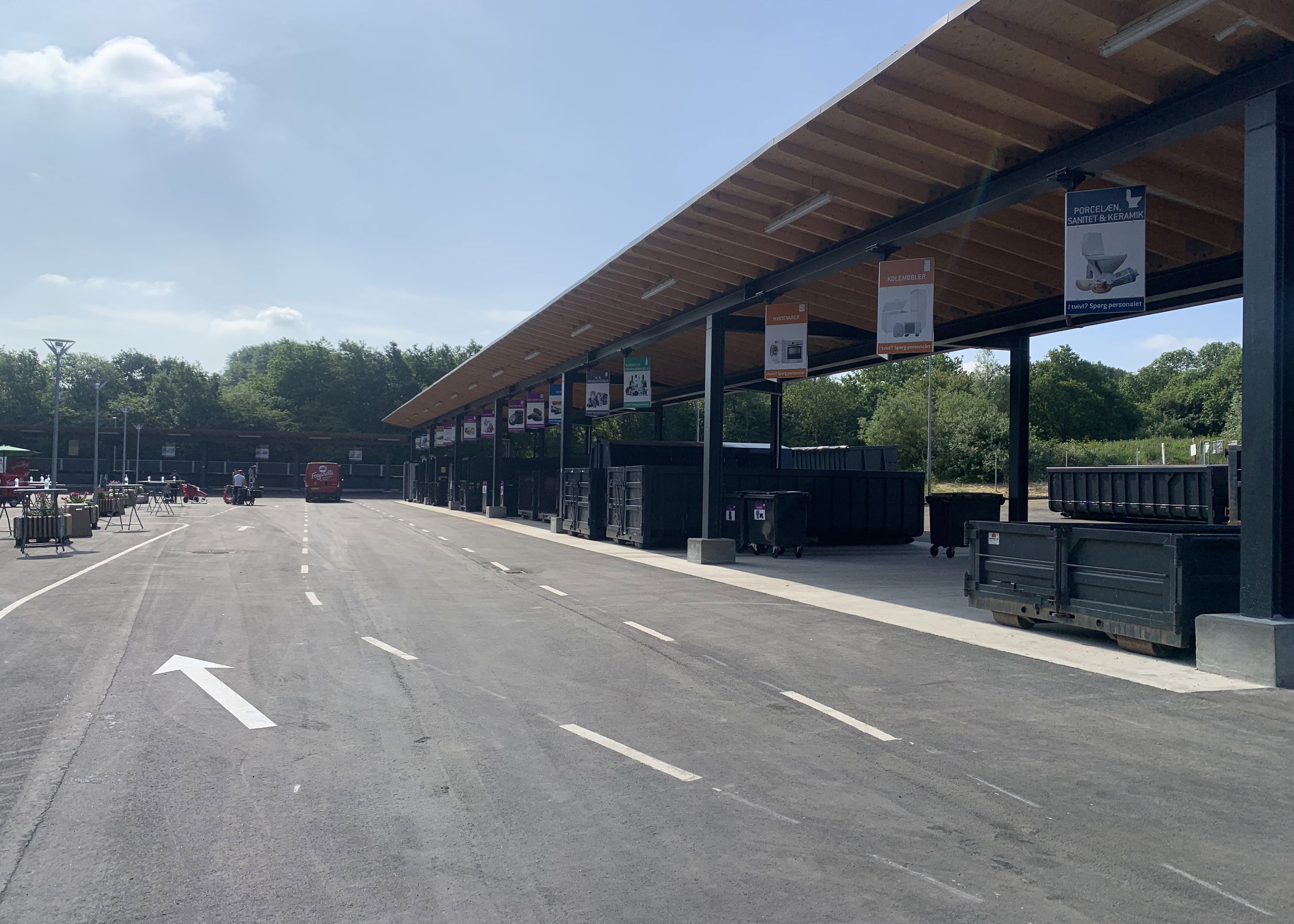 Børkop Genbrugsplads med en lang overdækket affaldssorteringsstation. Store containere er placeret under skiltede sektioner for forskellige affaldstyper. En asfalteret vej med kørselsanvisninger fører gennem pladsen, hvor en rød varevogn er synlig i baggrunden. Omkring pladsen er der grønne træer og en blå himmel med få skyer.