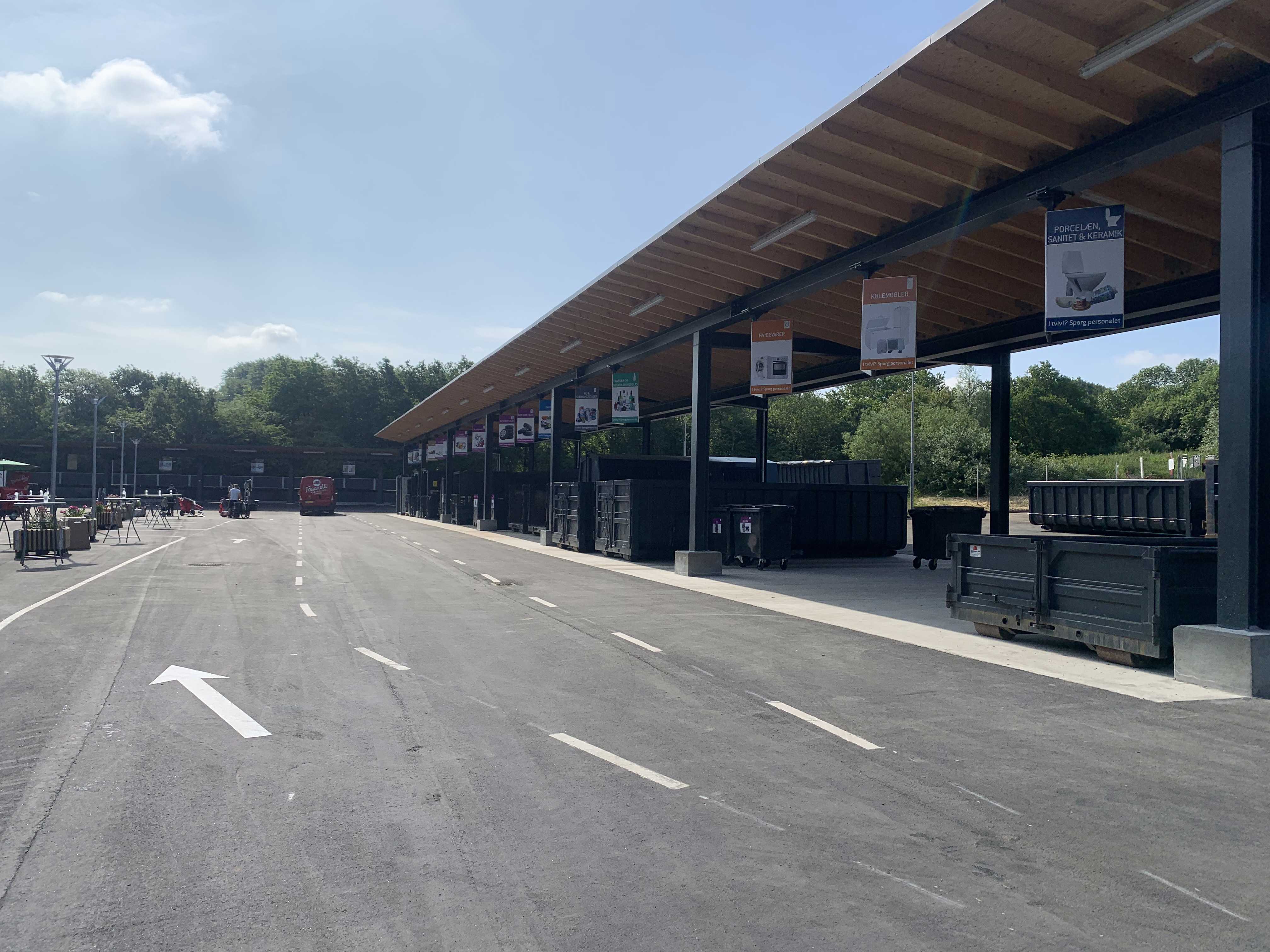 Børkop Genbrugsplads med en lang overdækket affaldssorteringsstation. Store containere er placeret under skiltede sektioner for forskellige affaldstyper. En asfalteret vej med kørselsanvisninger fører gennem pladsen, hvor en rød varevogn er synlig i baggrunden. Omkring pladsen er der grønne træer og en blå himmel med få skyer.